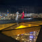 A couple walking on a sky deck with a glass floor at night, overlooking a brightly lit cityscape.