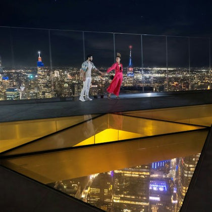 A couple walking on a sky deck with a glass floor at night, overlooking a brightly lit cityscape.
