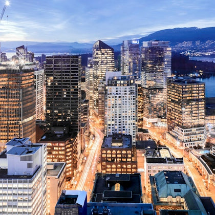 A bustling cityscape with illuminated tall buildings at dusk, showcasing a lively urban atmosphere and distant mountains.