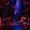 Performers in a brightly lit circus tent execute an aerial act with hoops and fabric under colorful lights.