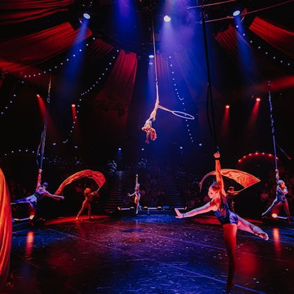 Performers in a brightly lit circus tent execute an aerial act with hoops and fabric under colorful lights.