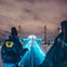 Two people walking on a lit-up structure at night with city lights and a cloudy sky in the background.