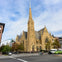 Large stone church with a tall spire on a city street corner, surrounded by parked cars and pedestrians.