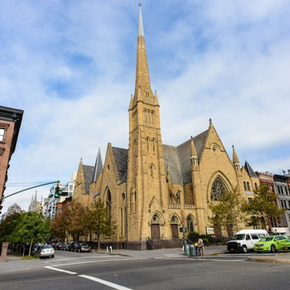 Large stone church with a tall spire on a city street corner, surrounded by parked cars and pedestrians.