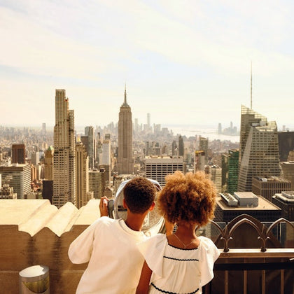 Two children look at a city skyline with tall buildings, including the Empire State Building, under a sunny sky.