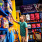 A boy in a green shirt reaches for a toy on a store shelf filled with colorful toys and backpacks. A sign reads, "No one is too tough for a soft blankie!"
