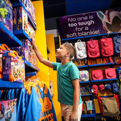 A boy in a green shirt reaches for a toy on a store shelf filled with colorful toys and backpacks. A sign reads, "No one is too tough for a soft blankie!"