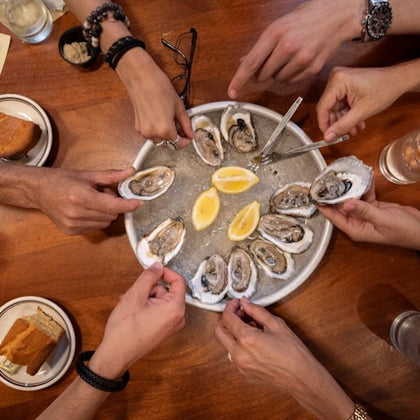Several hands holding oysters from a plate containing oysters and lemon wedges on a wooden table.