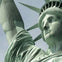 Close-up of the Statue of Liberty's face, crown, and raised arm against a light, cloudy sky.