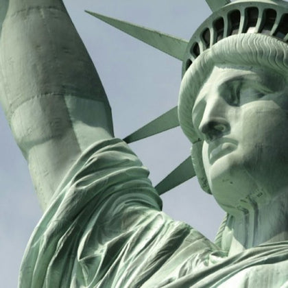 Close-up of the Statue of Liberty's face, crown, and raised arm against a light, cloudy sky.