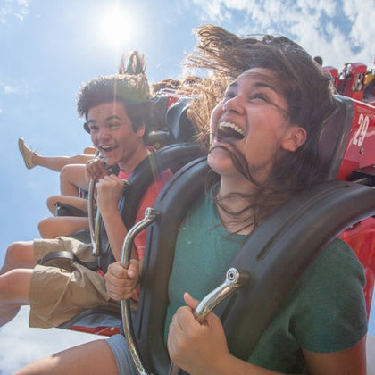 Roller coaster with red tracks and riders, surrounded by green trees, under a blue sky with scattered clouds.