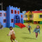 Children playing in an indoor play area with colorful structures, including a blue castle and yellow houses on green flooring.