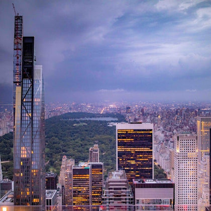 Aerial view of a cityscape at dusk, featuring tall buildings and a large park in the center under a cloudy sky.