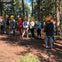 A group of people wearing safety equipment and orange helmets stand in a forest clearing, likely preparing for an activity.