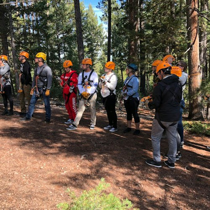 A group of people wearing safety equipment and orange helmets stand in a forest clearing, likely preparing for an activity.