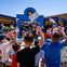 A group of children wave checkered flags at an outdoor event near a colorful building on a sunny day.