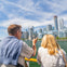 Two people on a boat looking at a city skyline with tall buildings under a blue sky with clouds.