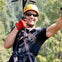 Person zip-lining through a forest, wearing a helmet and safety harness, smiling and gesturing with one arm.