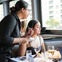 A woman seated at a table looks toward a server holding a notepad in a restaurant with large windows in the background.
