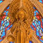 Carved wooden angel playing a trumpet, surrounded by colorful stained glass patterns in a cathedral setting.
