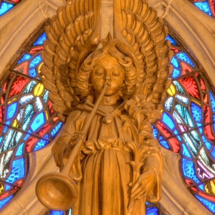 Carved wooden angel playing a trumpet, surrounded by colorful stained glass patterns in a cathedral setting.
