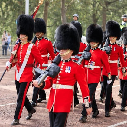 Guards in red uniforms and tall black hats march in formation, each holding a rifle, with a forest background.