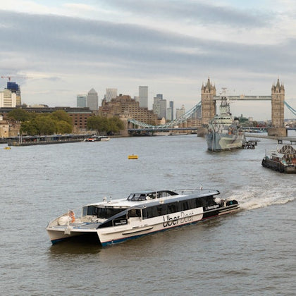 A river scene with a boat marked 'Uber Boat' cruising near Tower Bridge in London, with city buildings in the background.