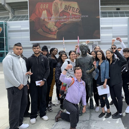 A group of people stands around a statue outdoors in front of a building with a large screen displaying sports memorabilia.