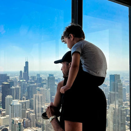 A man holds a young child on his shoulders while looking out from a tall building at a city skyline under a clear blue sky.