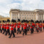 A marching band in red uniforms performs in front of Buckingham Palace under a partly cloudy sky.