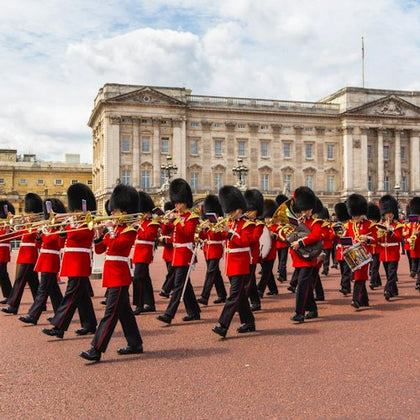 A marching band in red uniforms performs in front of Buckingham Palace under a partly cloudy sky.