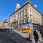 A busy city street intersection with a historic building housing retail stores, yellow taxis, and pedestrians in sunlight.