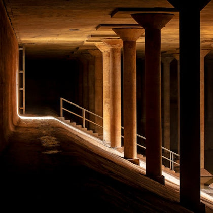 Dimly lit underground space with concrete columns, a sloped walkway, and metal railings illuminated by light.