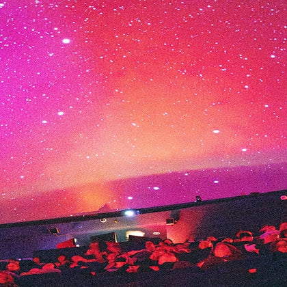 People seated and looking up at a planetarium dome displaying a vibrant, star-filled night sky with shades of red and purple.