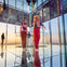 Two people in casual attire stand inside a mirrored observation space at sunset, with city lights visible in the reflection.