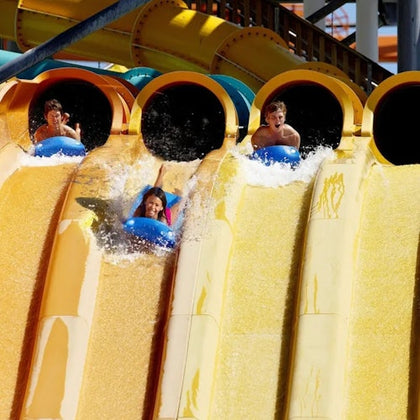Three people sliding down parallel water slides, each on an inflatable tube, with splashes of water around them.