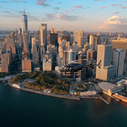 A helicopter flies over Lower Manhattan, New York City, with skyscrapers and the One World Trade Center in the background.