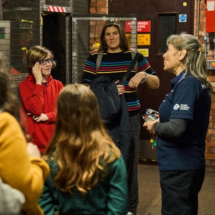 A group of people, including a guide in uniform, talking indoors near a brick wall and a door with safety signs.