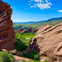 Red rock formations surround a lush green valley under a bright blue sky with scattered clouds.