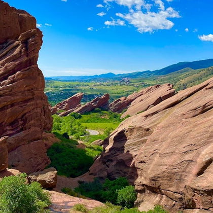 Red rock formations surround a lush green valley under a bright blue sky with scattered clouds.