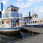 Two white and blue passenger boats docked at a pier, with a calm water surface and cloudy sky in the background.