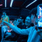A group of people laughing and enjoying a performance by a person dressed as a singer under blue stage lighting.
