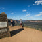Two people at a scenic lookout with a plaque and a railing, overlooking a mountain range under a clear sky.