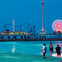 A beach scene at dusk with three people standing near the water, and an amusement park with rides visible in the background.