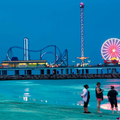 A beach scene at dusk with three people standing near the water, and an amusement park with rides visible in the background.