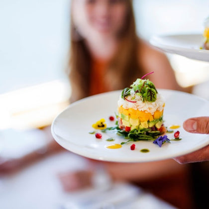A stacked dish of colorful vegetables and garnish on a white plate, with a blurred person in the background.
