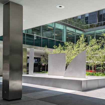 An outdoor plaza with modern metal sculptures, a metallic column, and glass-walled buildings in the background.