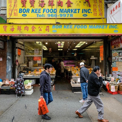People walk past the entrance of Bor Kee Food Market Inc., a brightly colored store selling various food items.