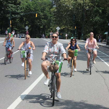 Five people riding bicycles on a tree-lined street, smiling and enjoying a sunny day.