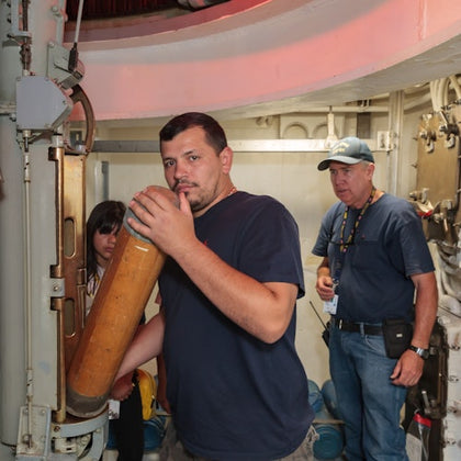 Three people are in a submarine interior. One man is holding a cylindrical object near a torpedo tube.